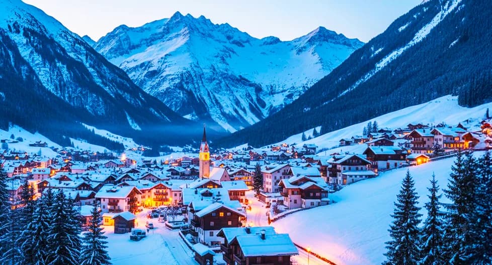 Austrian Alps ski resort village at dusk 2025 - traditional Tyrolean chalets and luxury ski hotels with warm evening lighting nestled in snow-covered valley, surrounded by dramatic mountain peaks and pristine ski slopes in winter
