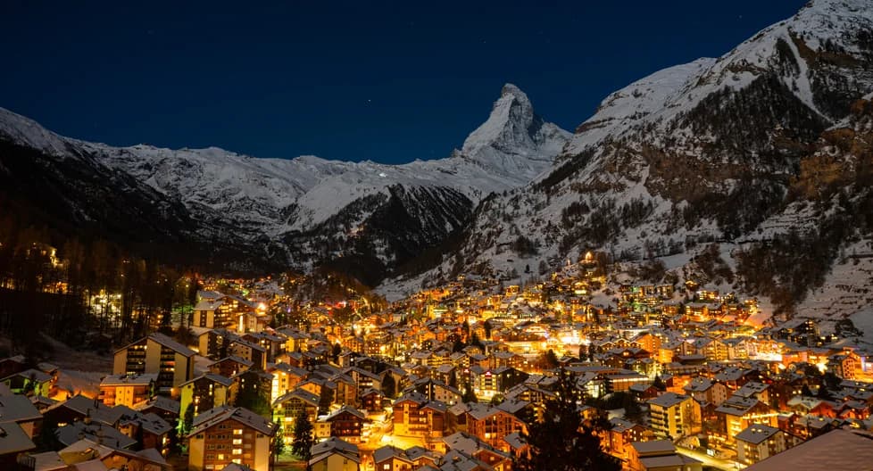 Zermatt Switzerland ski resort with iconic Matterhorn mountain at dusk - traditional wooden Swiss chalets and luxury alpine hotels with warm lighting, snow-covered slopes, car-free village, best Swiss ski destination winter 2025