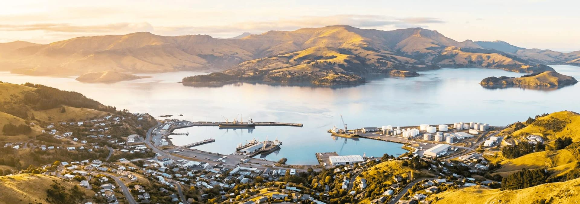 Aerial panoramic view of Lyttelton Harbor and Port Hills in Christchurch New Zealand at golden hour - volcanic crater harbor with working port, residential hillside suburbs, and Canterbury Plains with Southern Alps in background