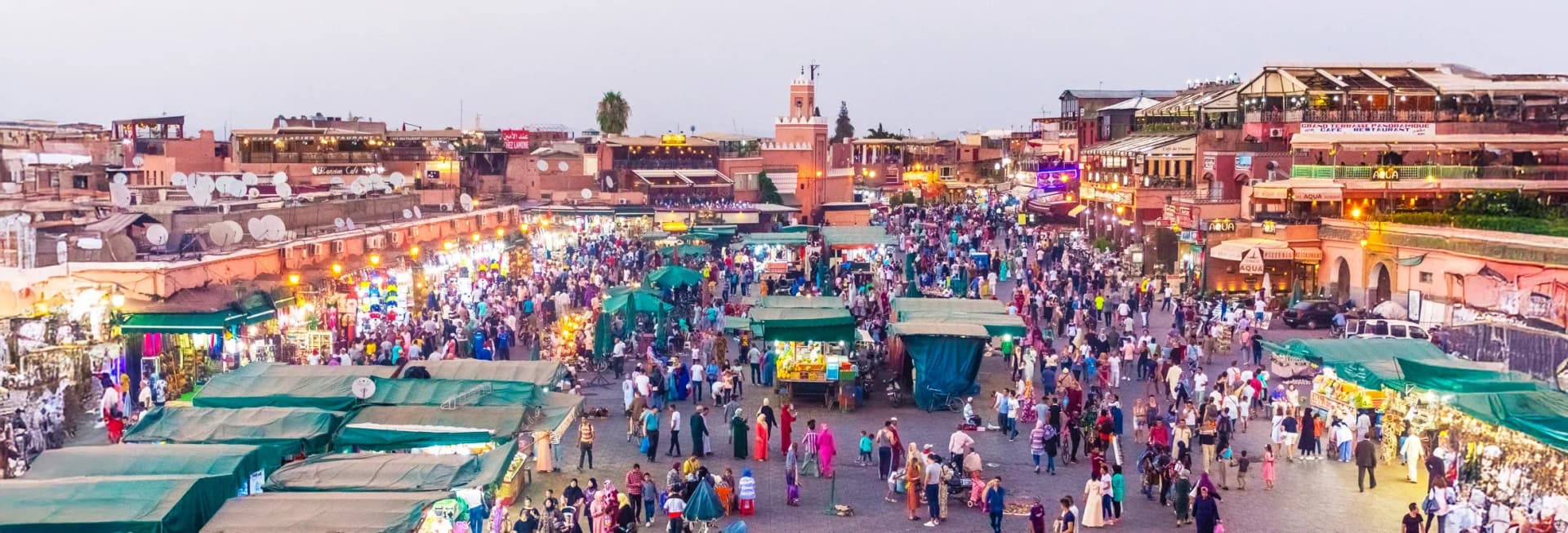Marrakech Morocco in December 2025 featuring the historic Koutoubia Mosque minaret tower rising above bustling Jemaa el-Fnaa square with vibrant souks, traditional Moroccan architecture, snow-capped Atlas Mountains in background, and palm trees under blue skies, showcasing perfect winter weather for exploring ancient medina markets, riads, and Sahara Desert excursions
