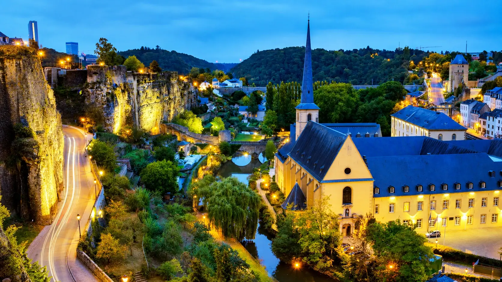 Panoramic view of Luxembourg City for our 2026 travel guide showing the UNESCO World Heritage old town perched above the Alzette River valley, with the historic Grund neighborhood, medieval stone fortifications, and Chemin de la Corniche walkway visible at golden hour