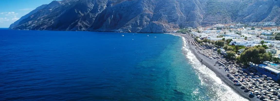Kamari Beach Santorini 2026 - famous black volcanic sand beach with organized sunbeds, beachfront restaurants and bars, Mesa Vouno mountain backdrop, clear blue Aegean waters, and family-friendly atmosphere